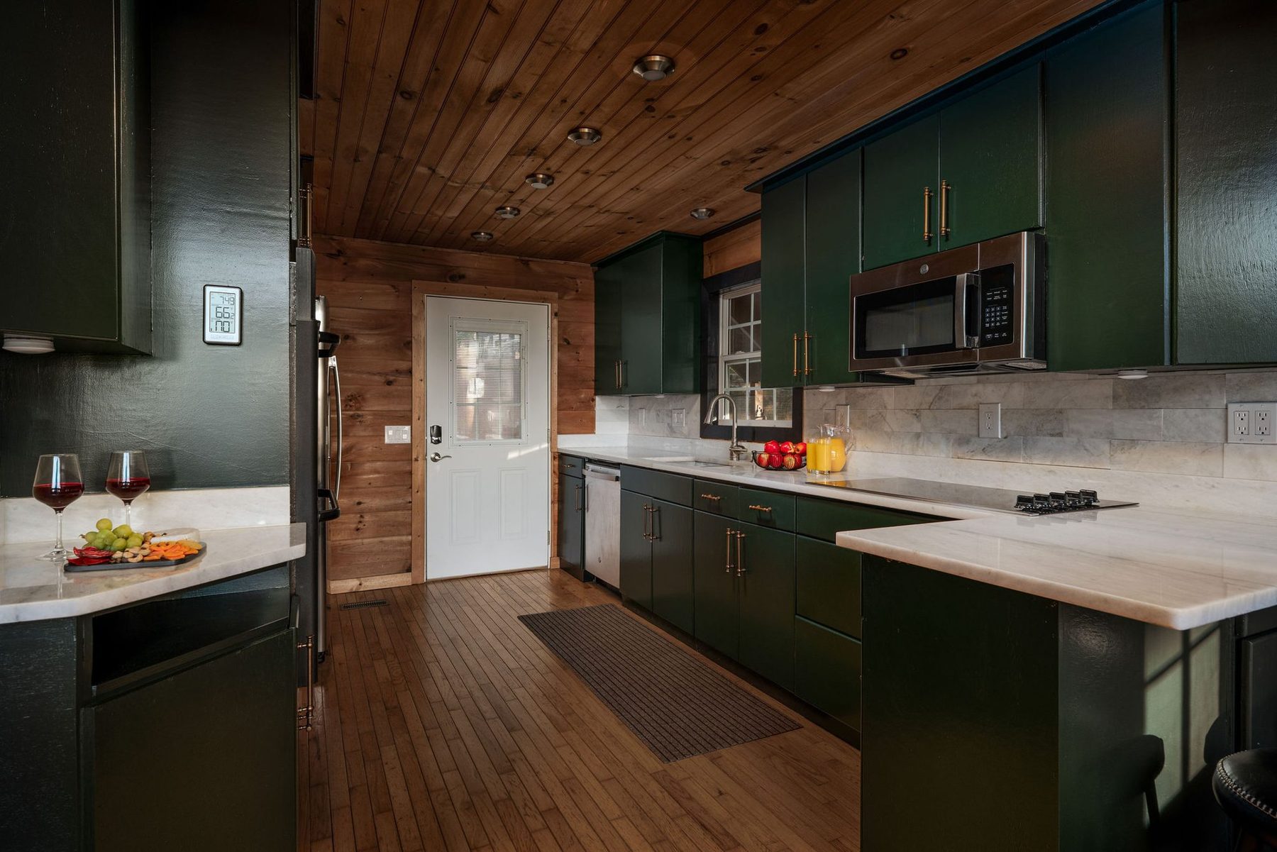 Kitchen with deep green cabinetry, white stone worktops and brass pulls against knotty pine