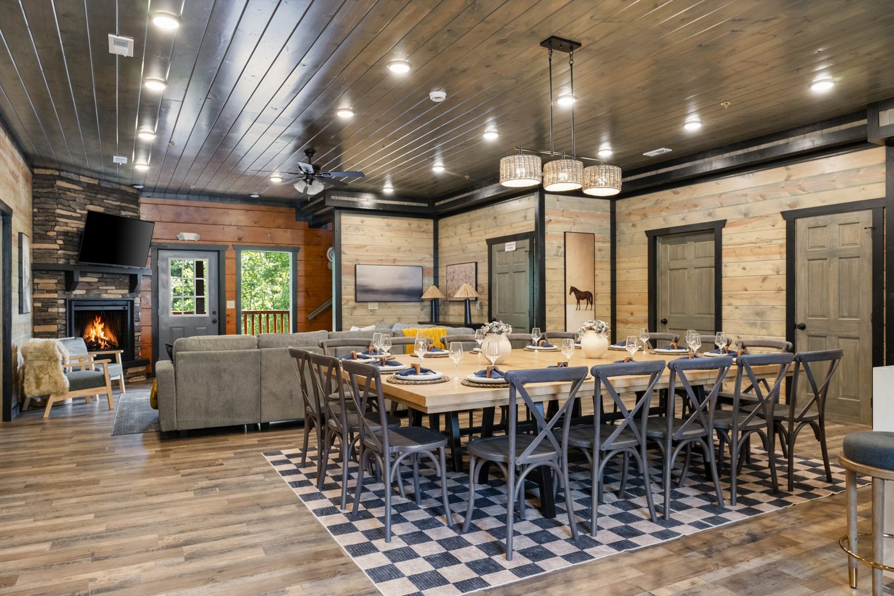 Great room under a dark plank ceiling: dining for twenty on a checked rug, stone fireplace beyond