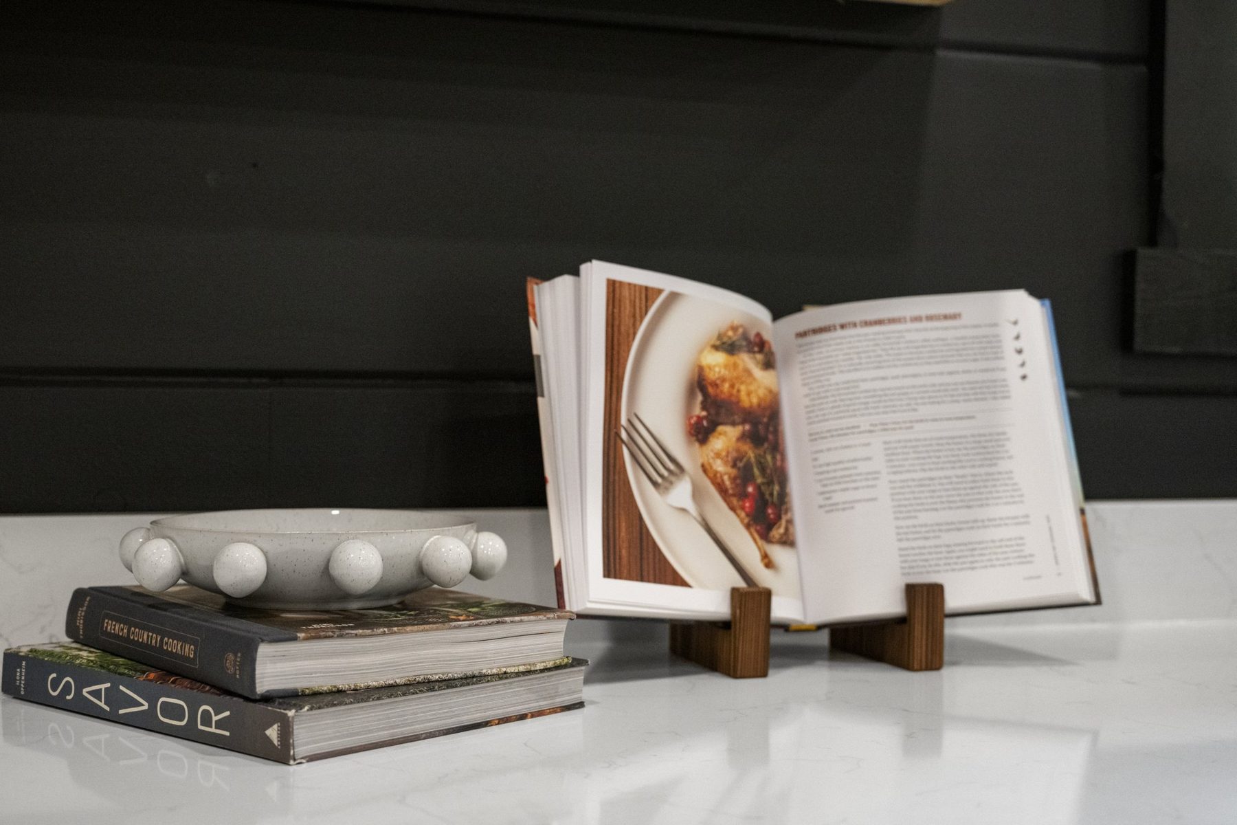 Open cookbook on a stand with stacked books on the white quartz counter, black cabinetry behind
