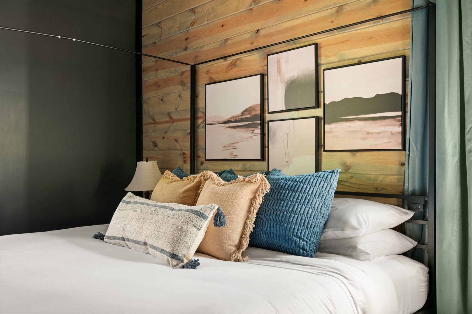 Bed dressed in blue and ochre cushions beneath a gallery of soft landscape prints