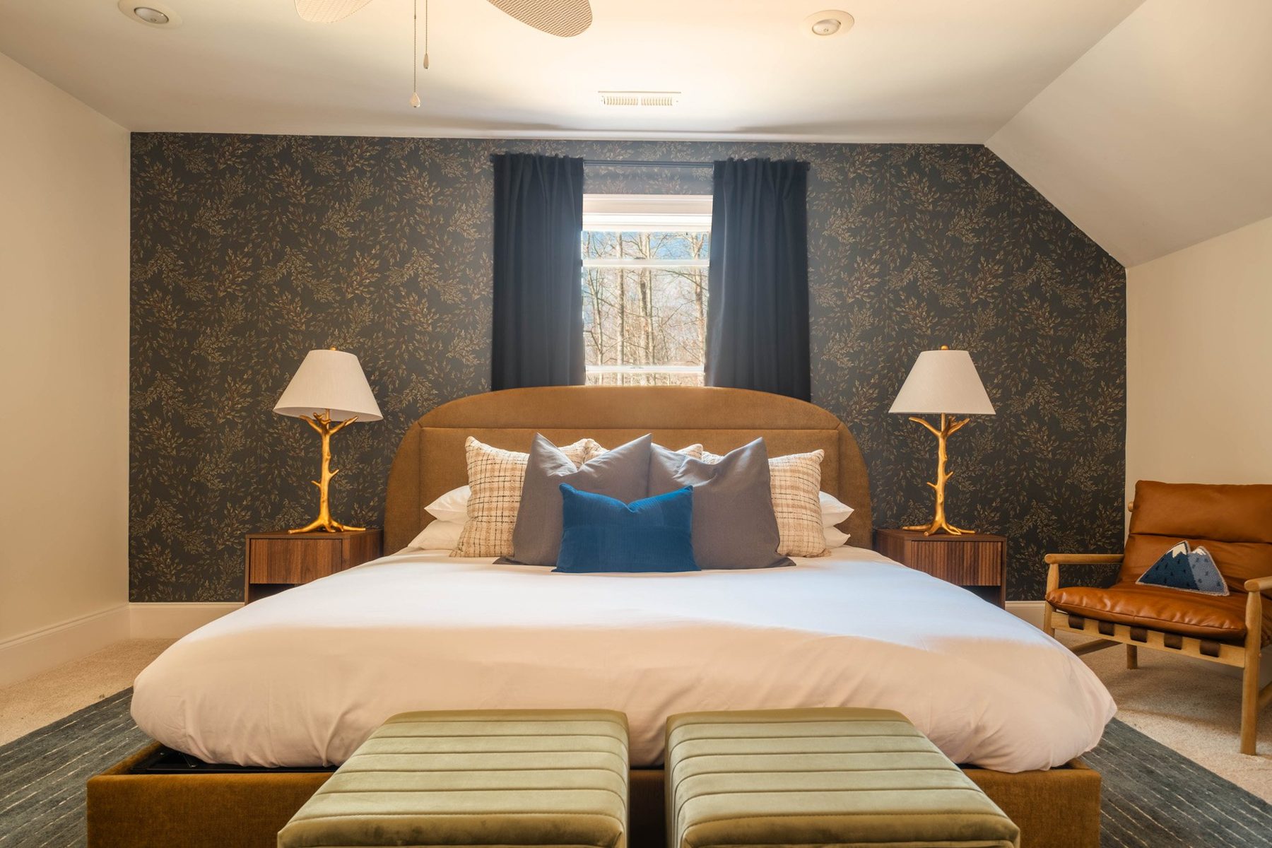 Bedroom with a dark botanical wallpaper wall, gold lamps and green velvet stools